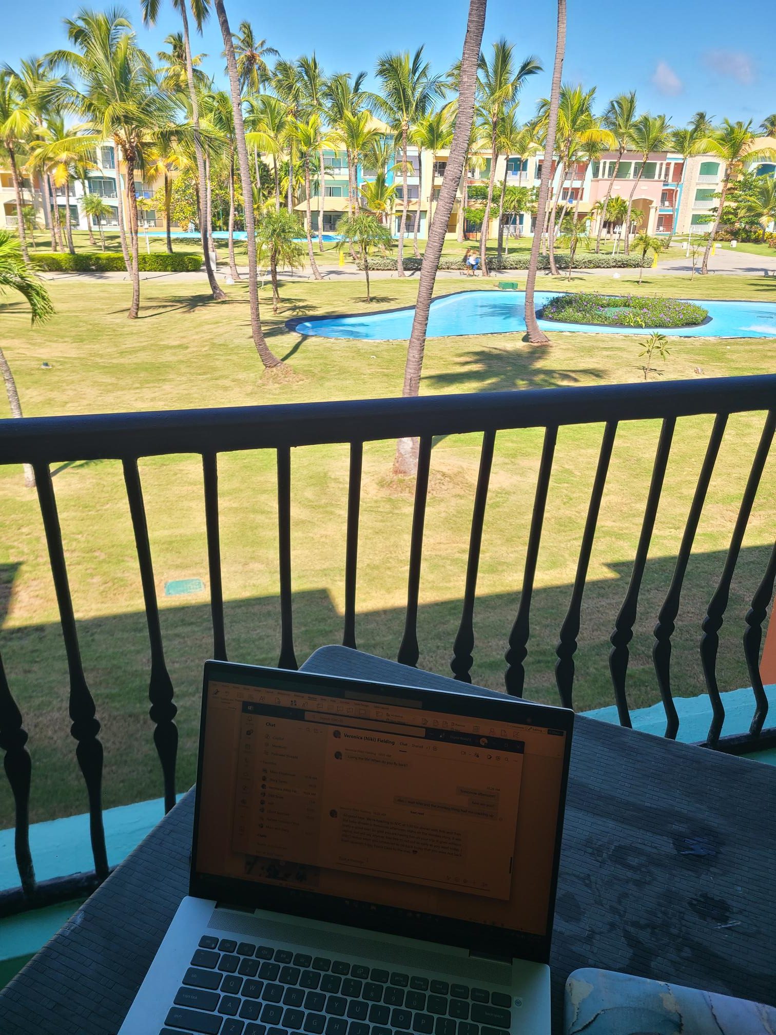 a laptop on a table set on a balcony overlooking palm trees and a fountain.